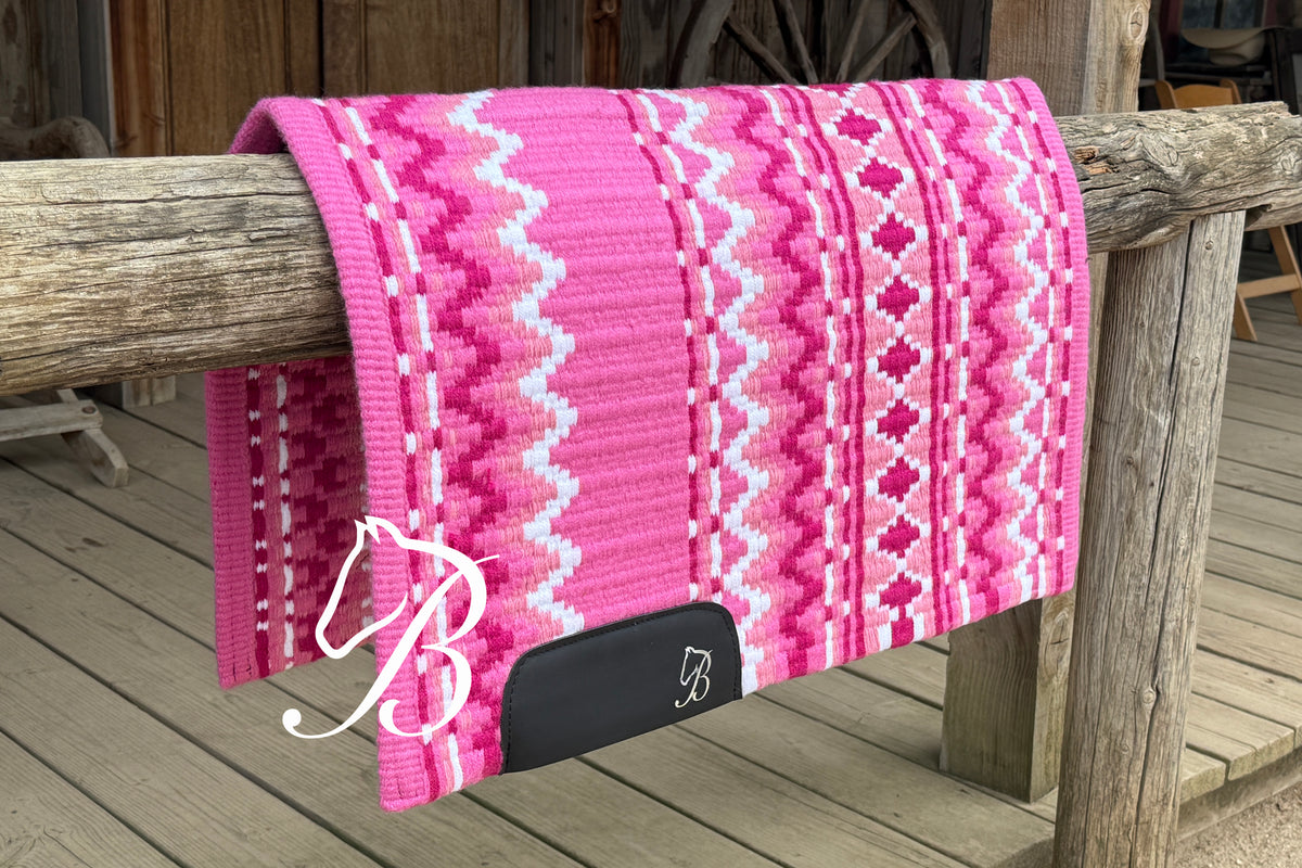 A pink patterned knit blanket draped over a rustic wooden fence post on a wooden deck. The blanket features a geometric zigzag and diamond pattern in shades of pink and white, with a black end tab and a white embroidered monogram logo.
